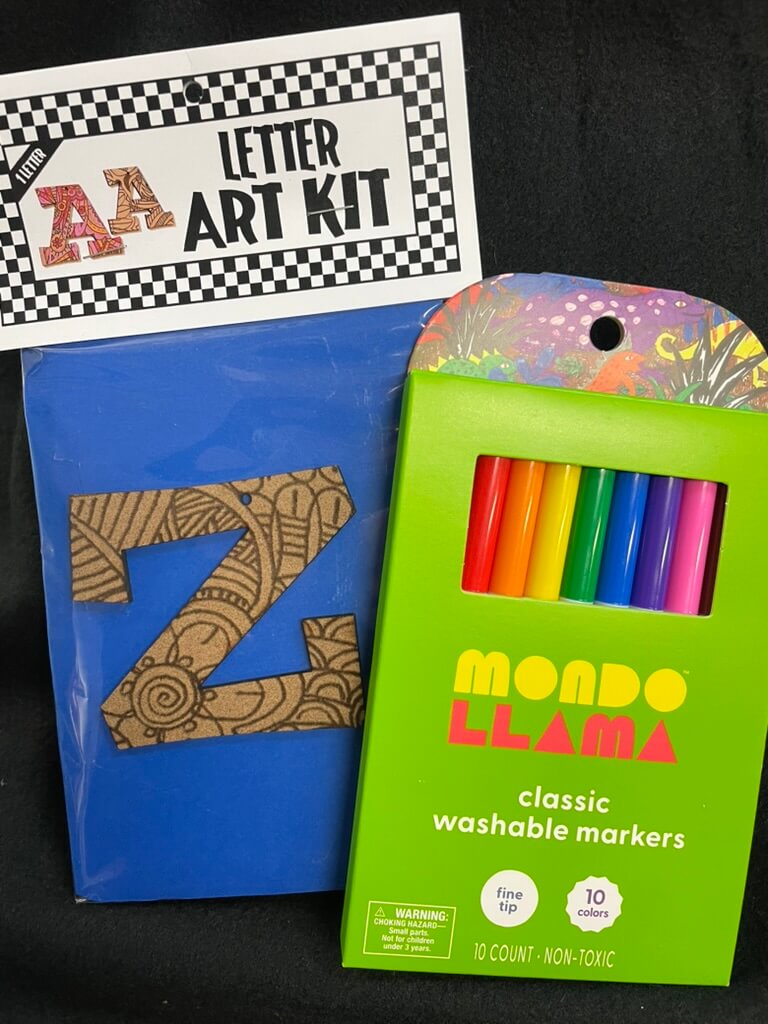 The image shows a part of an art kit with a letter 'Z' shaped template and a set of colorful washable markers. The packaging is predominantly green with a colorful design and text that reads 'A to Z Art Kit by Mando Lama'.