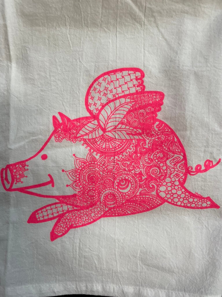 A tea towel with an pink and white design featuring a flying pig.