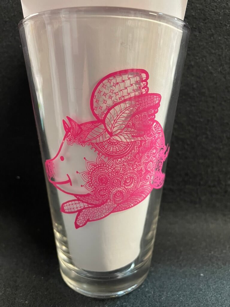 A clear pint glass with a pink illustration of a flying pig on it.
